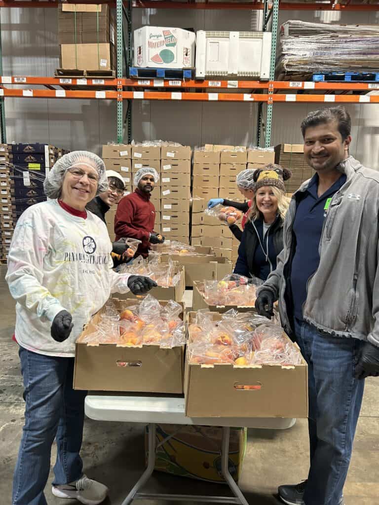 Five people smiling and packing food into plastic bags at a table inside a warehouse filled with boxes, wearing gloves and hairnets.