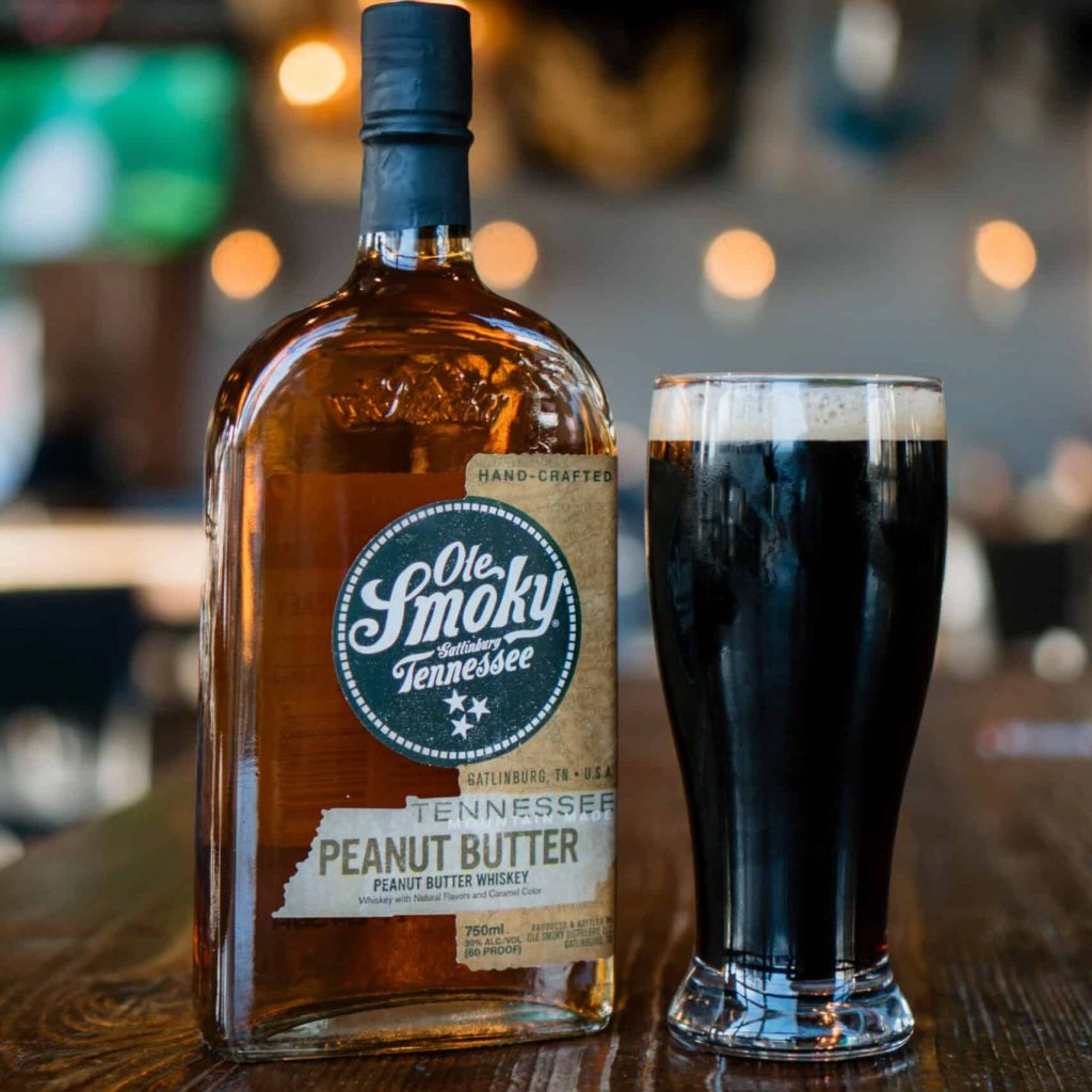 A bottle of Ole Smoky Tennessee Peanut Butter Whiskey sits beside a tall glass of dark beer on a wooden bar with a blurred background of lights and decorations.