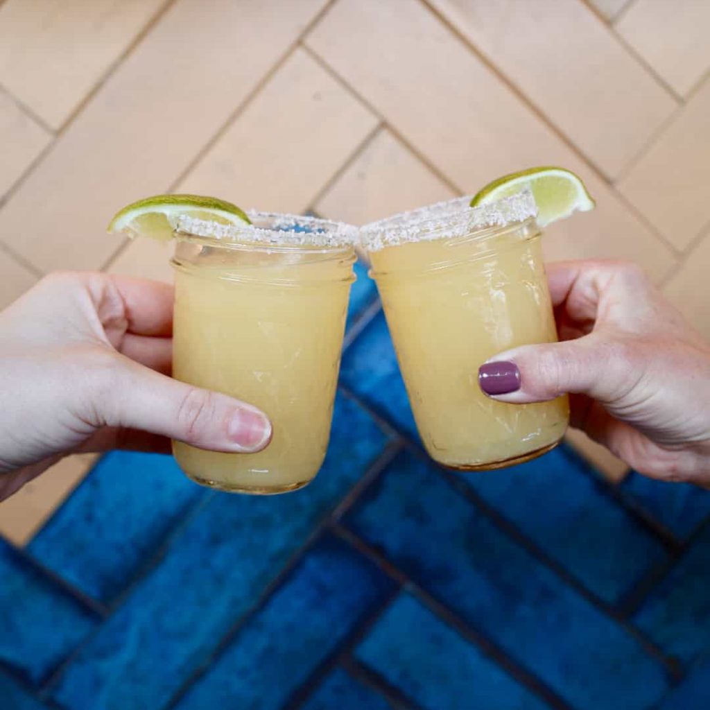 Two hands holding up mason jar glasses filled with a yellow cocktail, garnished with lime wedges and a salted rim, as if making a toast. The background features herringbone-patterned wood and blue tile flooring.